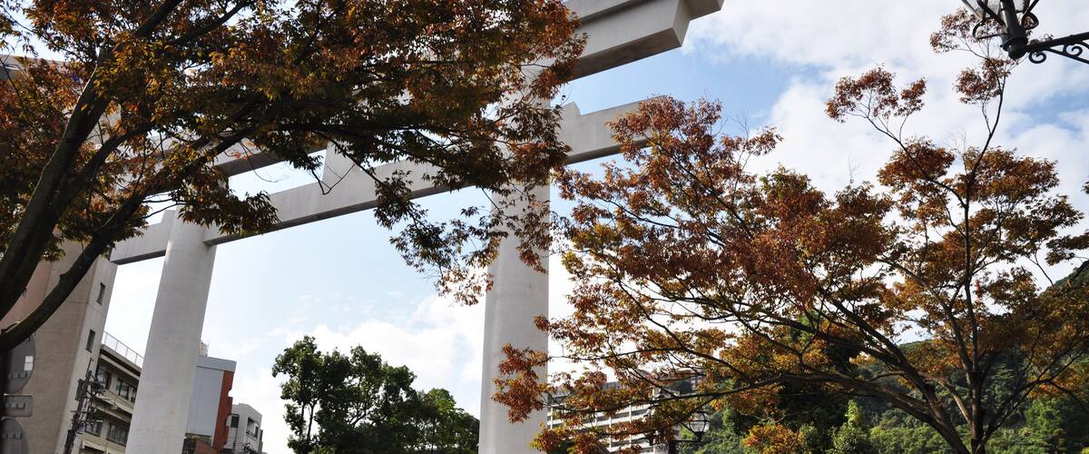 A large stone torii gate in a park with autumn trees and modern buildings in the background.