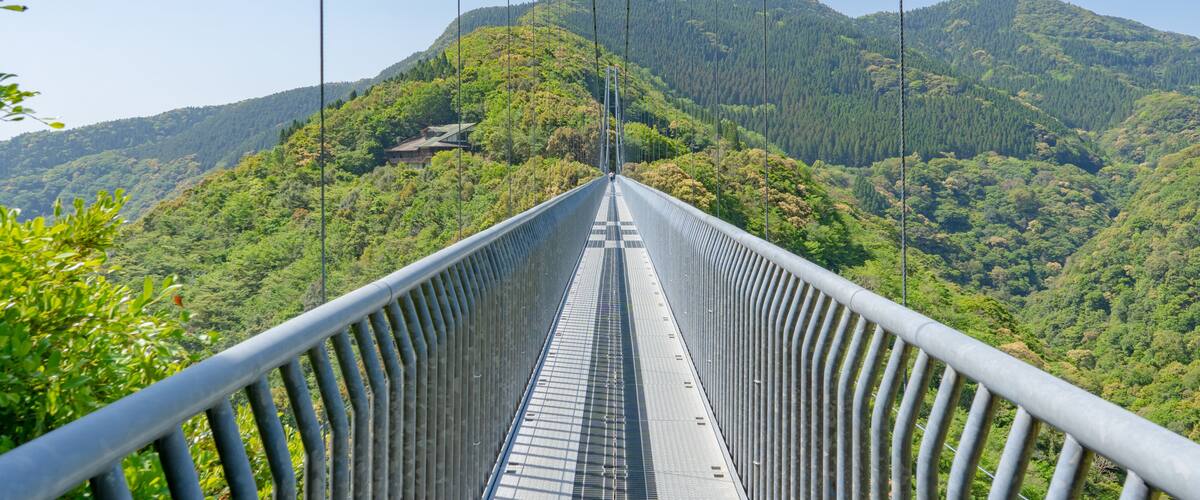 A suspension bridge with a metal railing, surrounded by lush green hills and a clear sky.