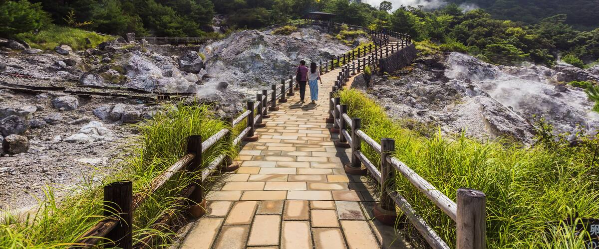 A stone pathway leading uphill with wooden railings, surrounded by lush greenery and a mountainous backdrop.