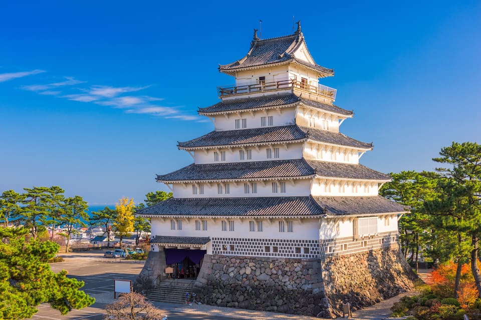 A multi-tiered traditional Japanese castle with a stone base, surrounded by trees and a clear sky.