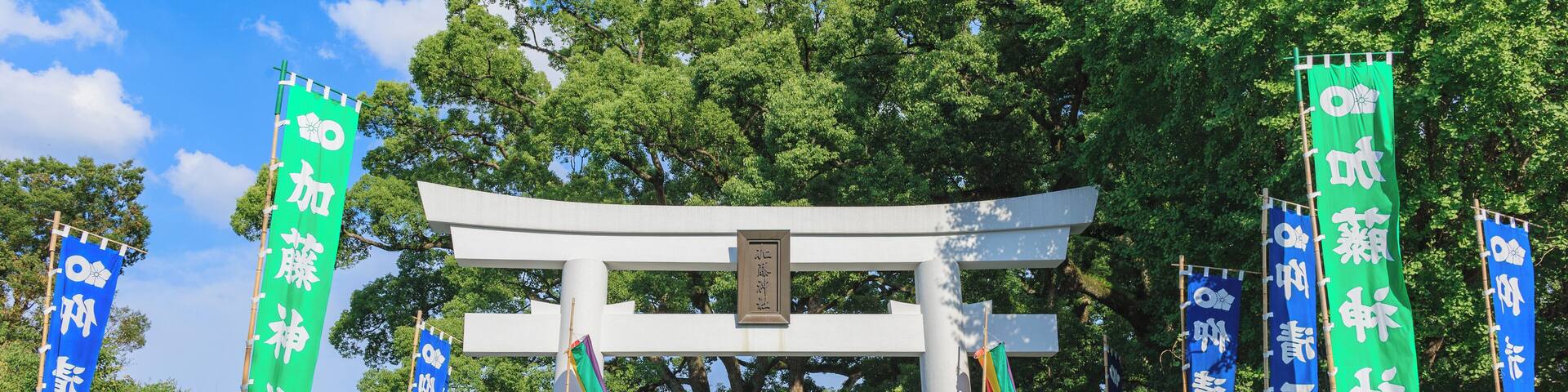 A traditional Japanese torii gate with green and blue banners, surrounded by trees and stone lanterns.