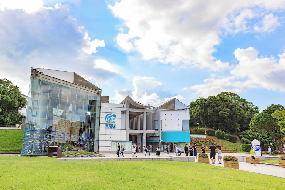 A modern building with glass facades and a blue sign, surrounded by green grass and trees.