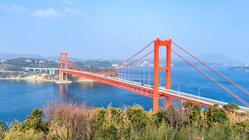 A red suspension bridge over a body of water with hills in the background.