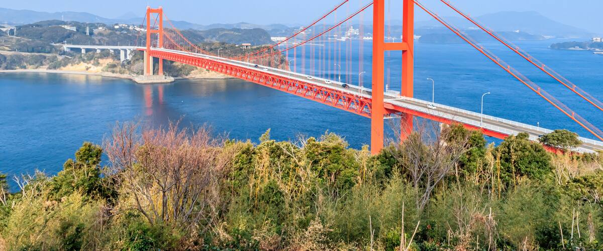 A red suspension bridge over a body of water with hills in the background.