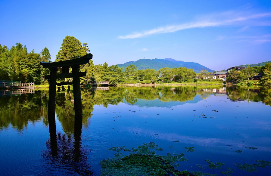 A serene lake with a traditional Japanese torii gate in the foreground, surrounded by lush greenery and mountains in the background.