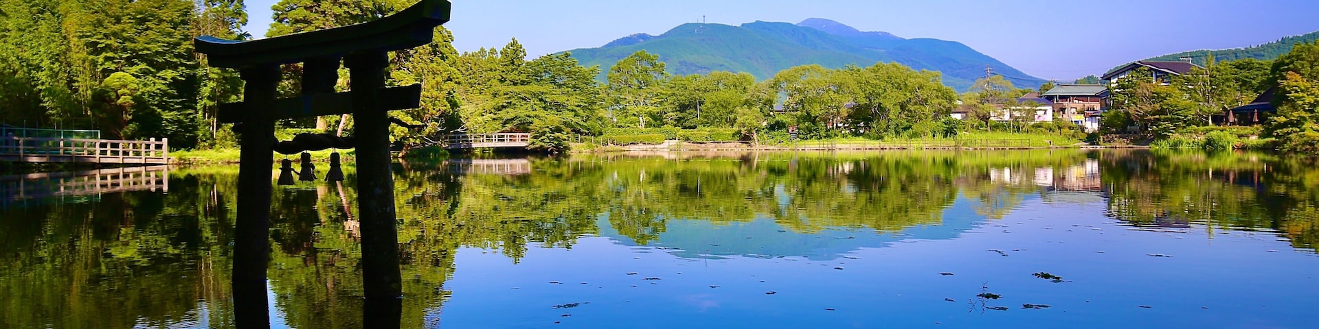 Um lago tranquilo com um tradicional portão torii japonês em primeiro plano, cercado por vegetação exuberante e montanhas ao fundo.
