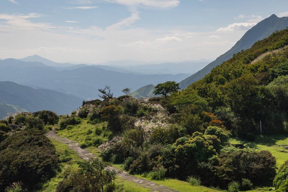 A mountainous landscape with a winding path, lush greenery, and distant mountain ranges under a clear sky.