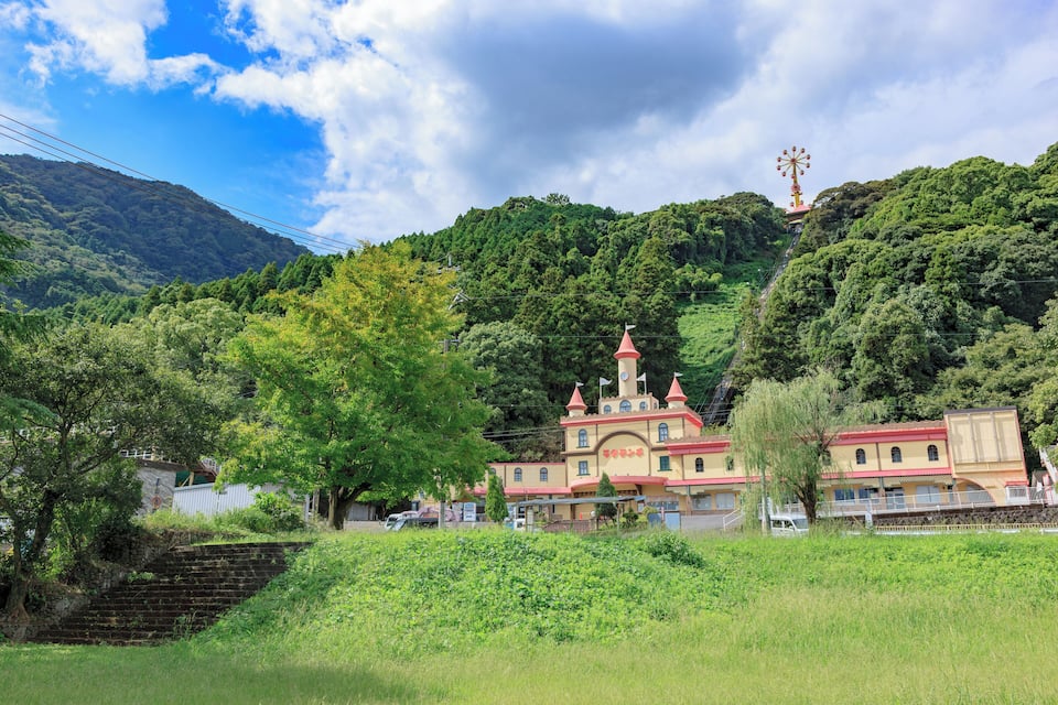 Un edificio con aspecto de castillo, con techo rojo y varias torres, rodeado de colinas verdes y árboles exuberantes.