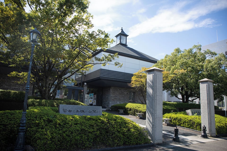 A traditional Japanese building with a stone facade, surrounded by lush greenery and a clear blue sky.
