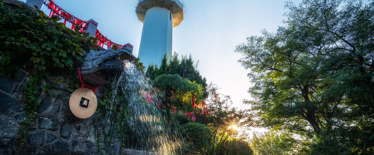 A lighthouse with a water feature, surrounded by greenery and stone walls.