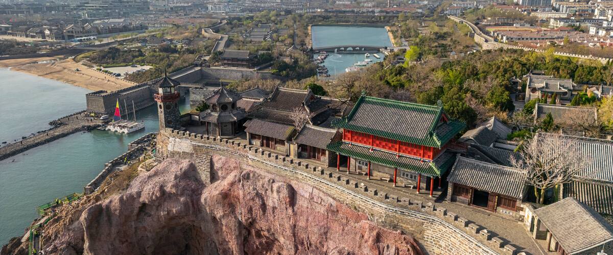 A traditional Chinese fortress with a green-roofed building, a rocky cliff, and a river in the background.
