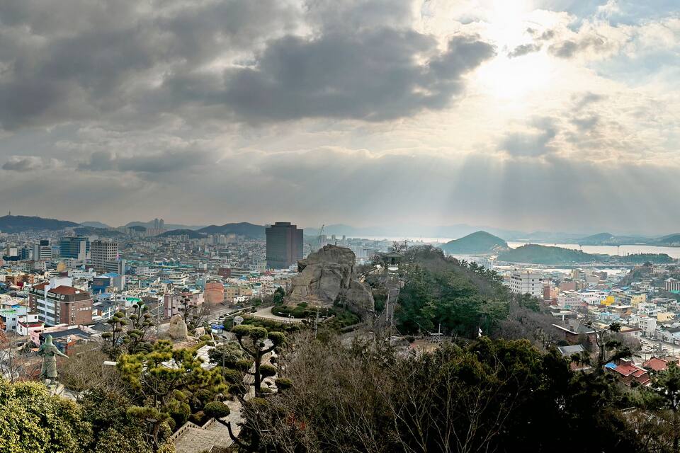 A cityscape with a prominent rock formation in the foreground, a dense urban area with various buildings, and a distant mountain range under a cloudy sky.