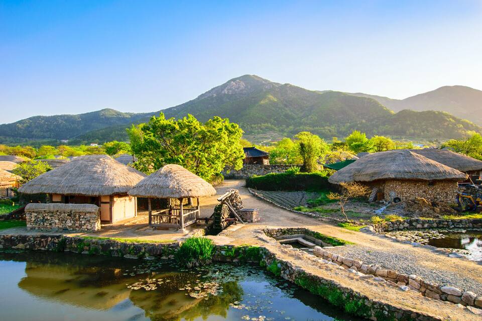 Traditional thatched-roof huts, a stone well, and a calm pond in a rural village setting with mountains in the background.