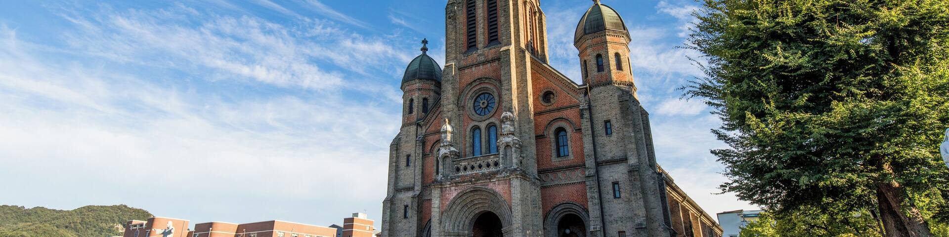 Una iglesia histórica con una destacada torre de campanas y dos cúpulas, rodeada por una plaza con gente y árboles.