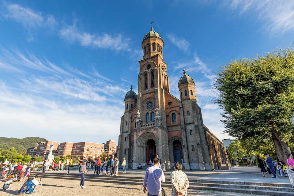 A historic church with a prominent bell tower and two domes, surrounded by a plaza with people and trees.