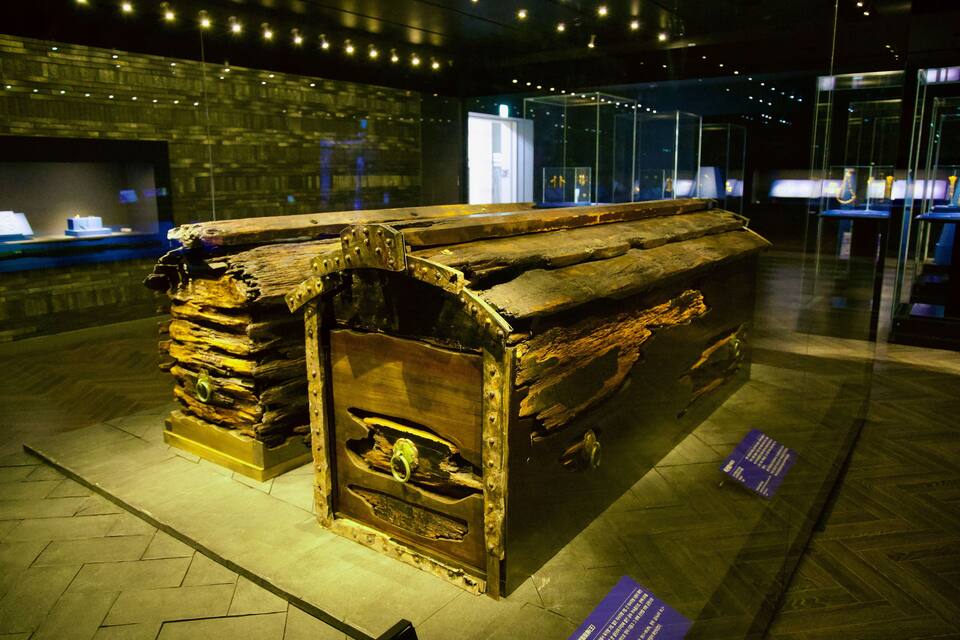 Un vieux coffre en bois renforcé de métal, exposé dans un musée, à l’intérieur de vitrines et accompagné de panneaux explicatifs.
