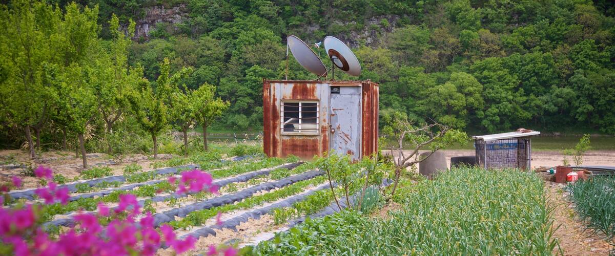 A rural landscape with a rusted metal structure, satellite dishes, and rows of crops.
