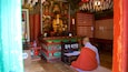 Een monnik in traditionele kleding zittend in een tempel met een Boeddhabeeld, wierookbrander en gebedsraden.