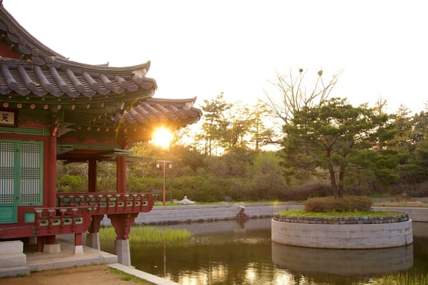 A traditional Korean pavilion with a tiled roof, a stone bridge, and a pond surrounded by trees.