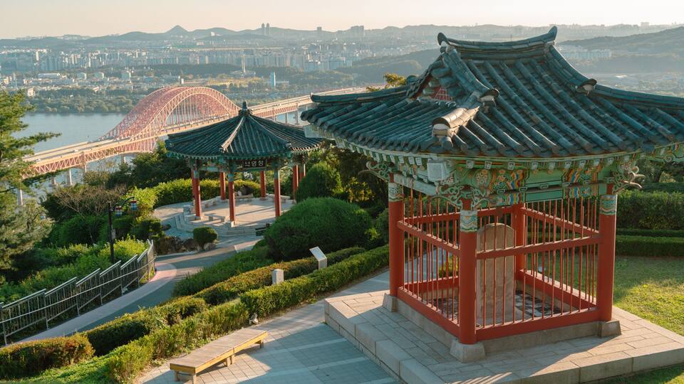 Traditional Korean architecture with a pavilion, a bridge, and a cityscape in the background.