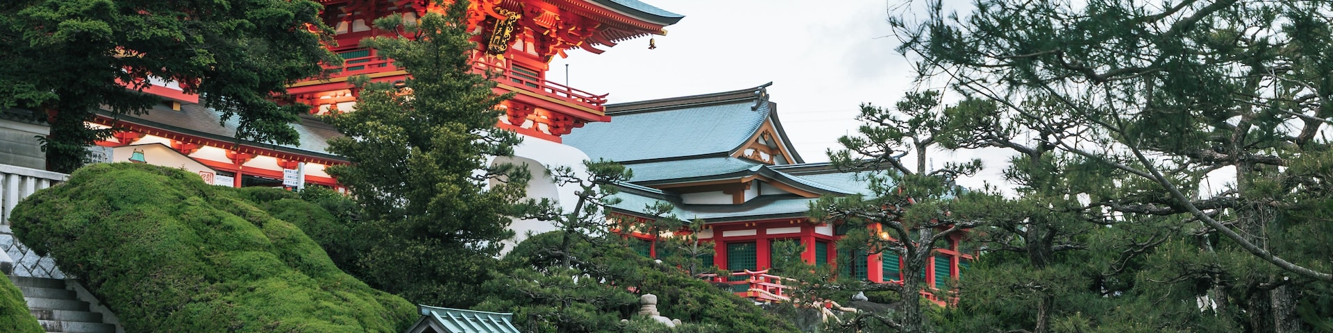 Um templo japonês tradicional com arquitetura vermelha e verde, rodeado por vegetação exuberante e uma escadaria de pedra.