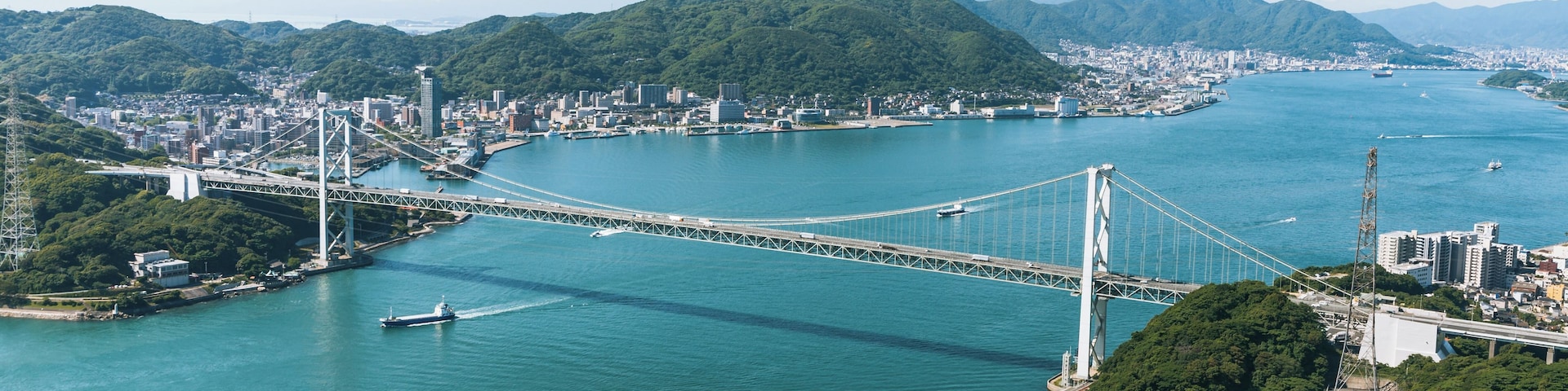 Uma ponte suspensa sobre um rio, rodeada por montanhas e um horizonte urbano.