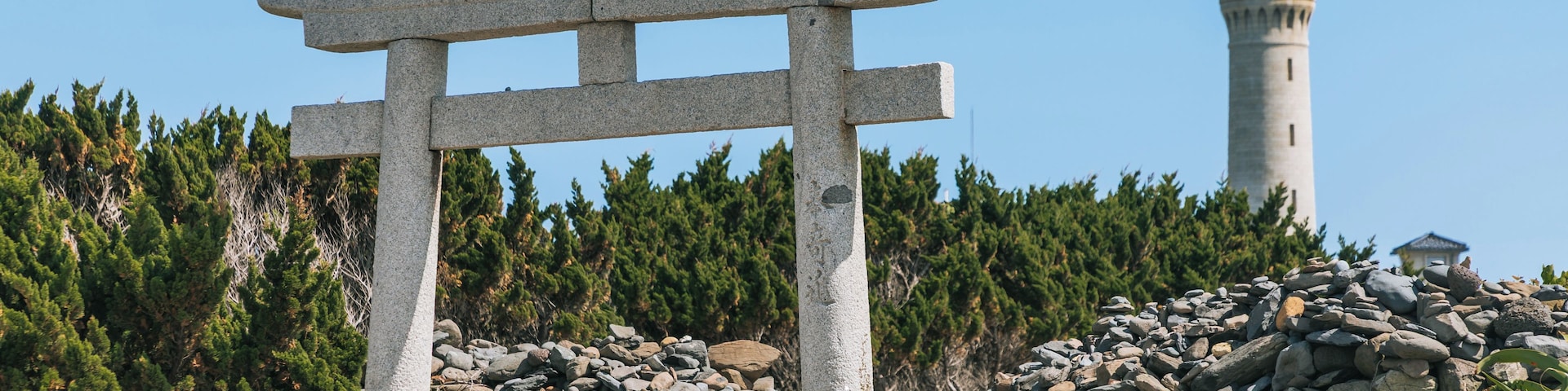 Um portal torii de pedra em um ambiente natural, com um farol ao fundo.