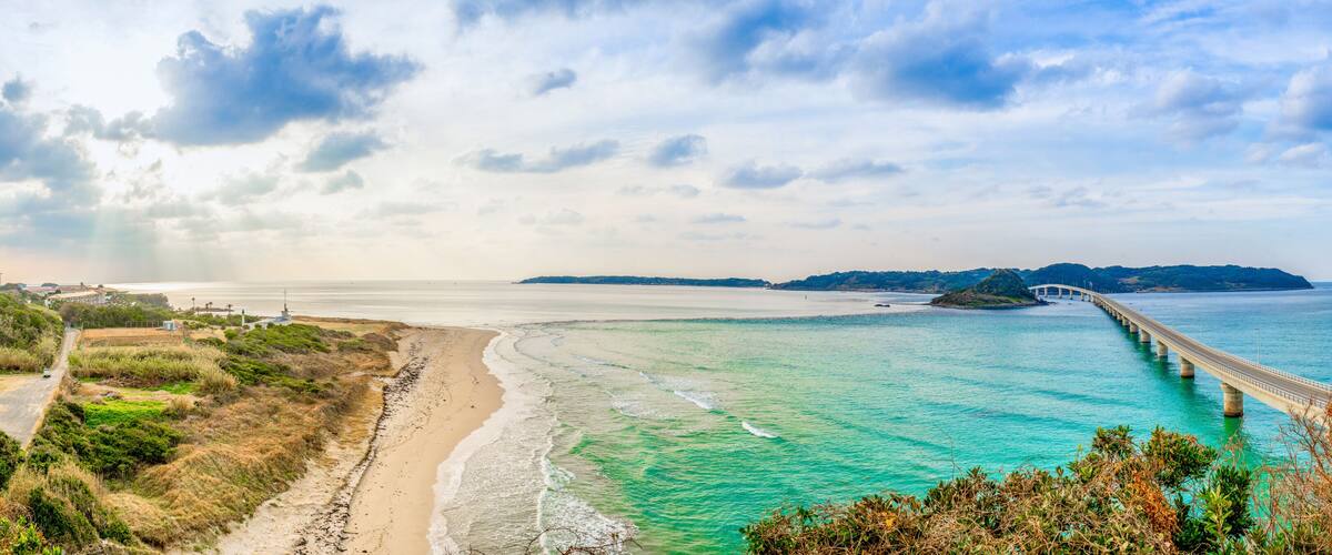 A coastal landscape with a beach, turquoise waters, a white bridge, and distant hills.