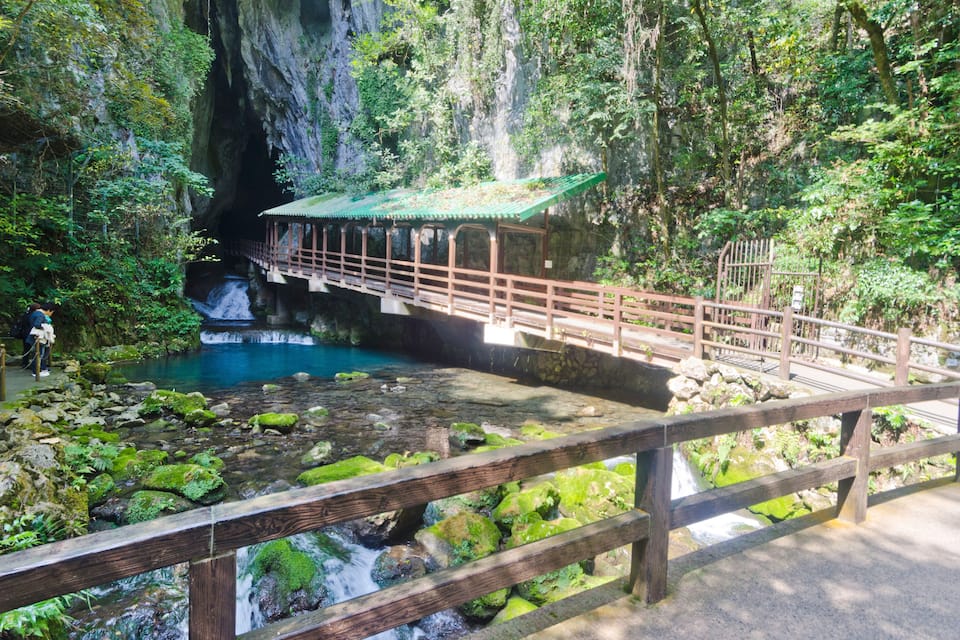 A wooden bridge over a clear blue stream with a traditional pavilion on the left.