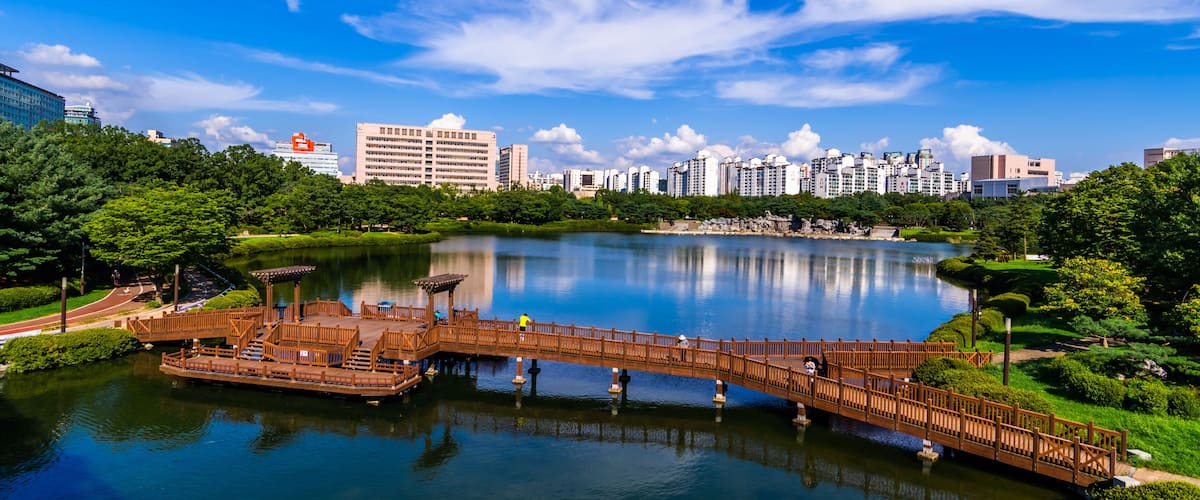 A serene lake with a wooden bridge, surrounded by lush greenery and modern high-rise buildings in the background.