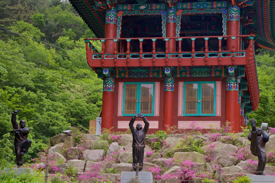 Ein traditionelles rotes Gebäude mit türkisfarbenen Akzenten, umgeben von Statuen und blühenden, rosa Pflanzen.
