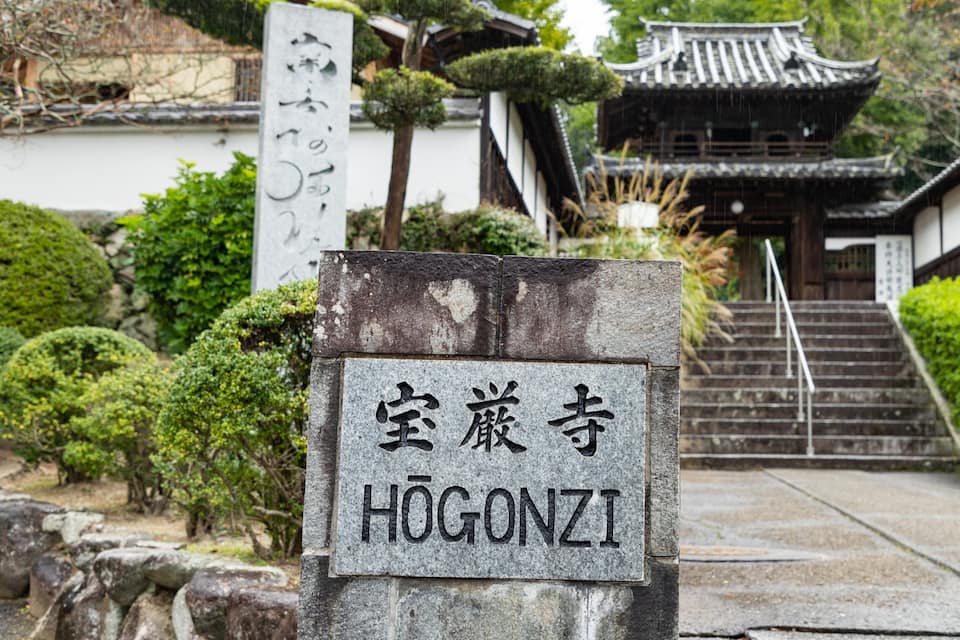A stone sign with Japanese characters and the English name "Hōgōnzi" in front of a traditional Japanese building with a tiled roof and stairs leading up to it.