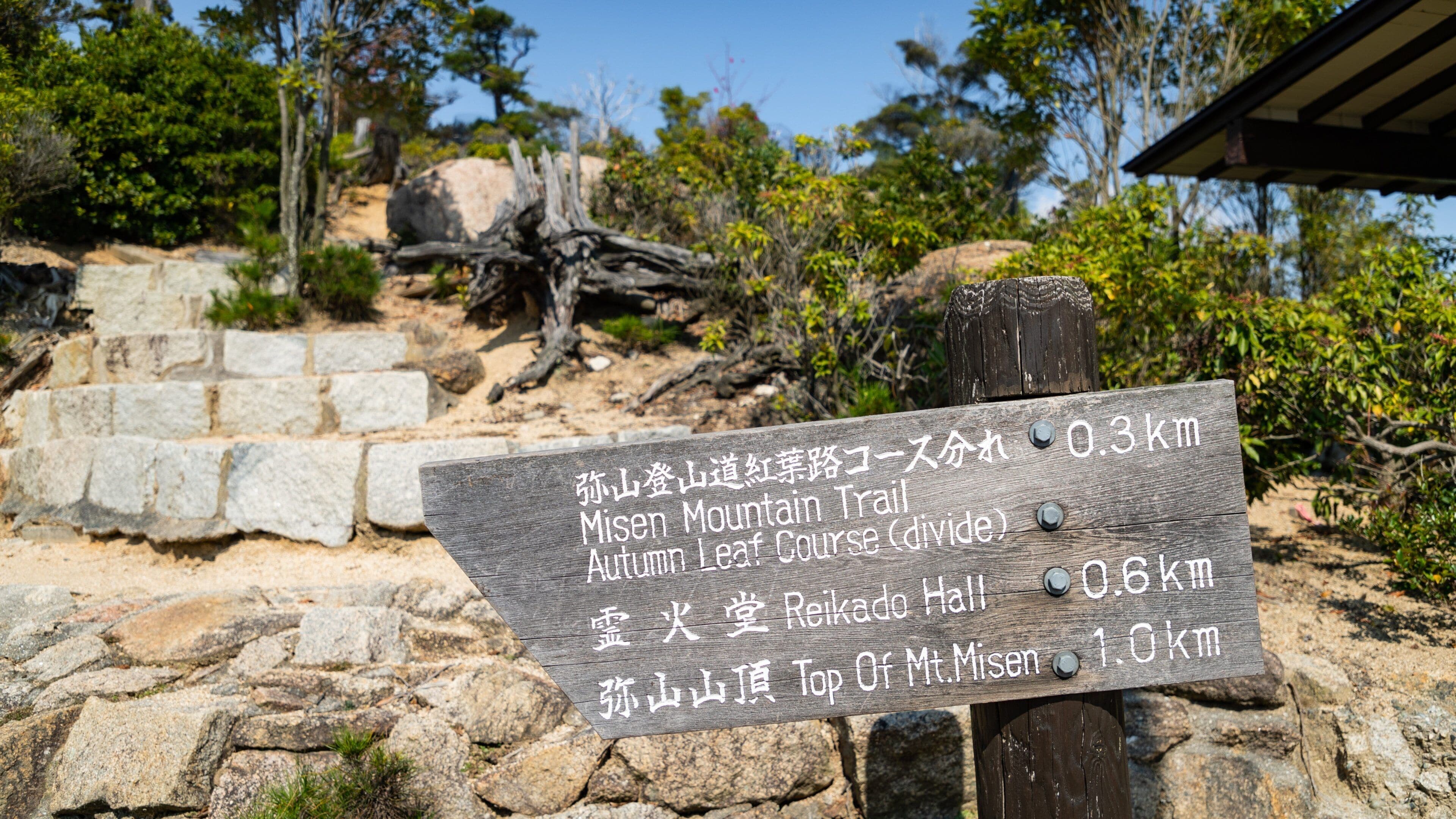 A wooden sign with Japanese text indicating distances to Misen Mountain Trail, Autumn Leaf Course, Reikado Hall, and Top of Mt. Misen.