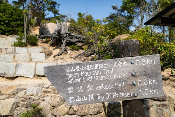 Ein hölzernes Schild mit japanischem Text, das die Entfernungen zum Misen-Berg-Pfad, dem Herbstlaub-Pfad, der Reikado-Halle und der Spitze des Berges Misen angibt.