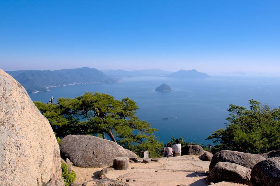 Eine gebirgige Landschaft mit freiem Blick auf das Meer, felsigem Gelände und einer Person, die auf einem Felsen sitzt.