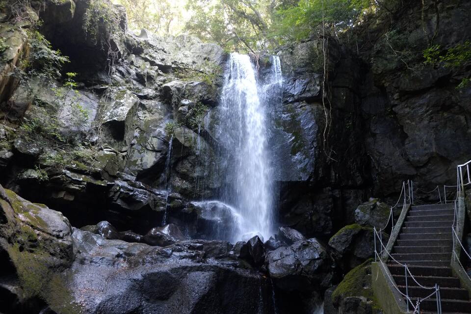 A waterfall cascading down a rocky cliff, surrounded by lush greenery and a staircase leading up to the water.
