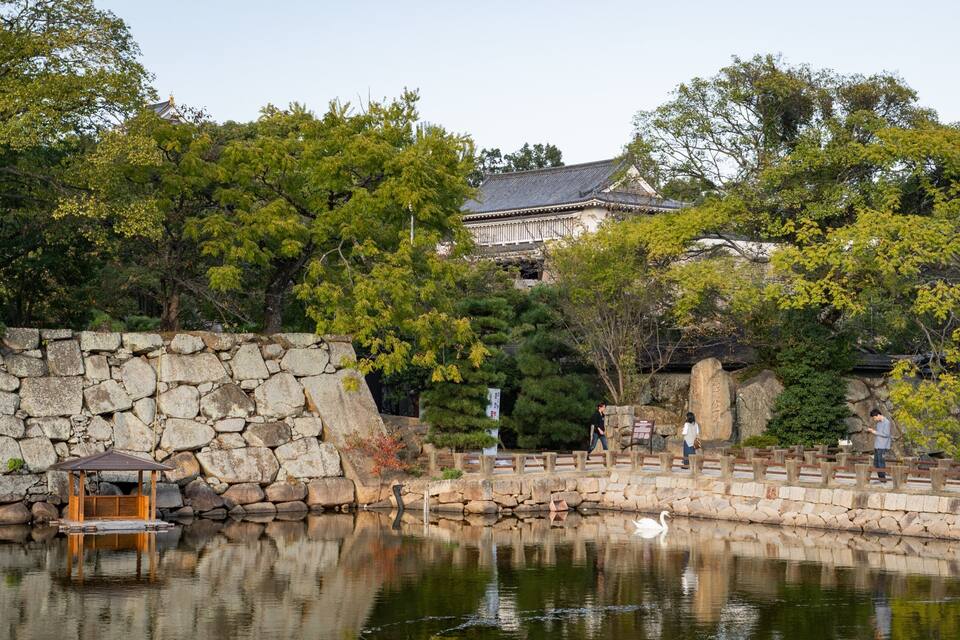 A traditional Japanese garden with a pond, stone wall, wooden bridge, and a building in the background.