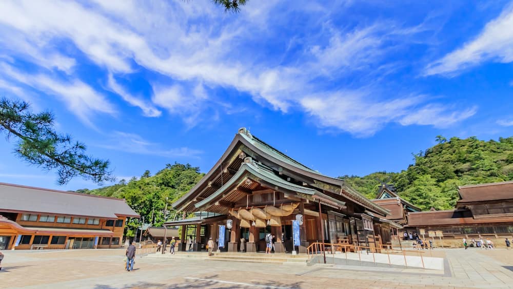 Ein traditionelles japanisches Gebäude mit einem Ziegeldach, umgeben von einem gepflasterten Bereich und Bäumen.