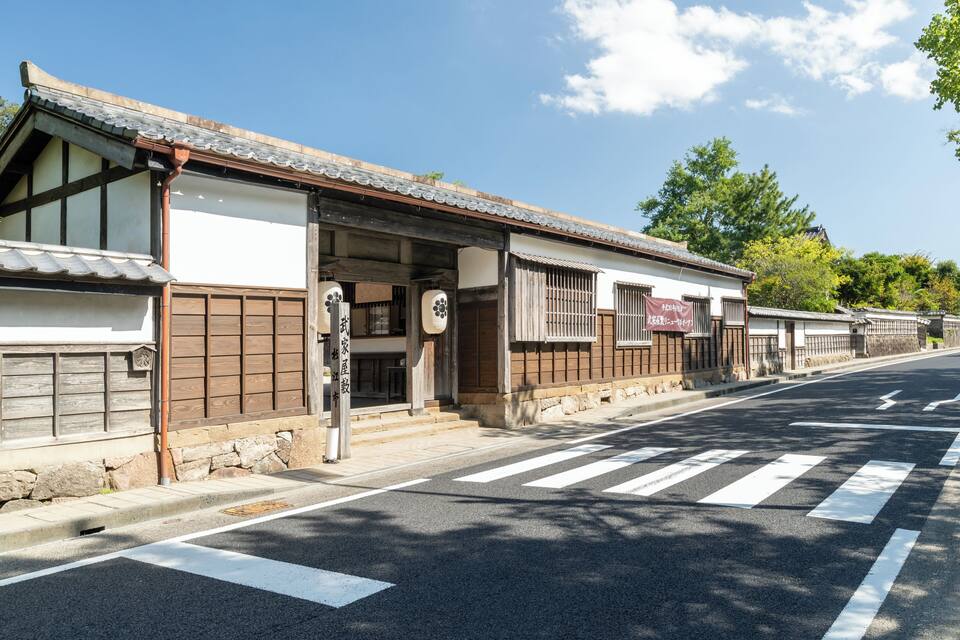 Un carrer tradicional japonès amb edificis de fusta, un pas de zebra i un cel blau clar.