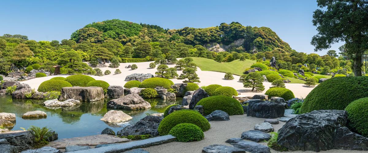 A serene Japanese garden with a pond, neatly trimmed bushes, and a backdrop of lush hills.