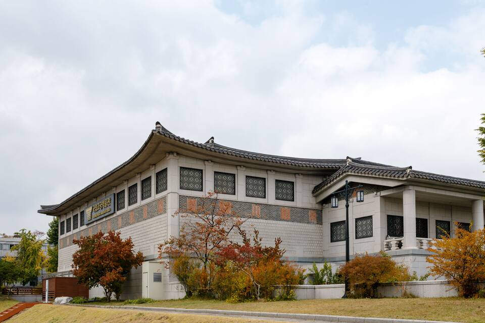 A traditional Korean building with a curved roof and tiled eaves, surrounded by autumn-colored trees.