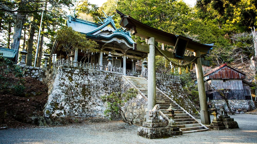 A traditional Japanese temple with a staircase leading up to a shrine, surrounded by trees and stone structures.