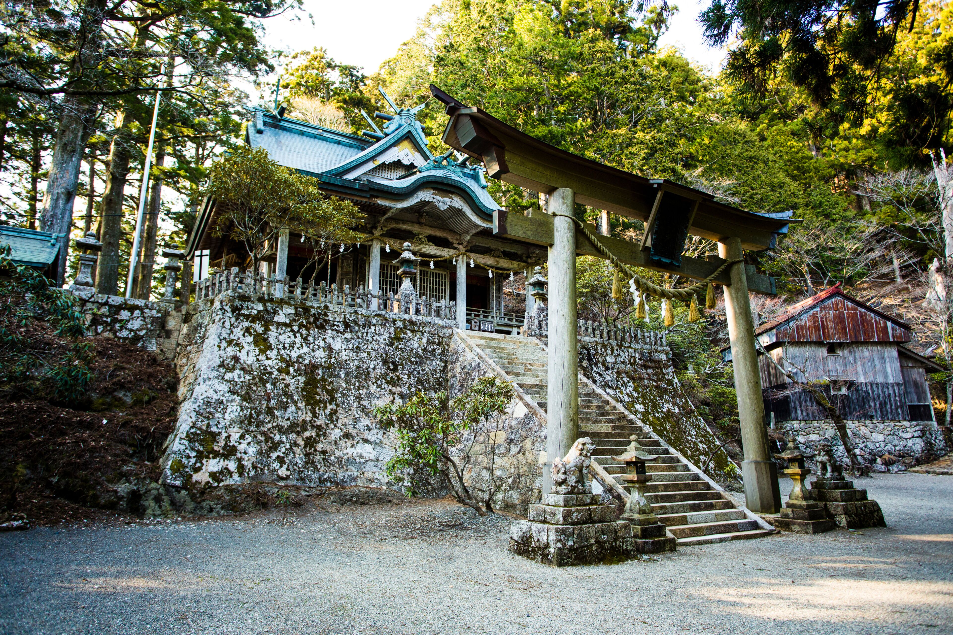 A traditional Japanese temple with a staircase leading up to a shrine, surrounded by trees and stone structures.