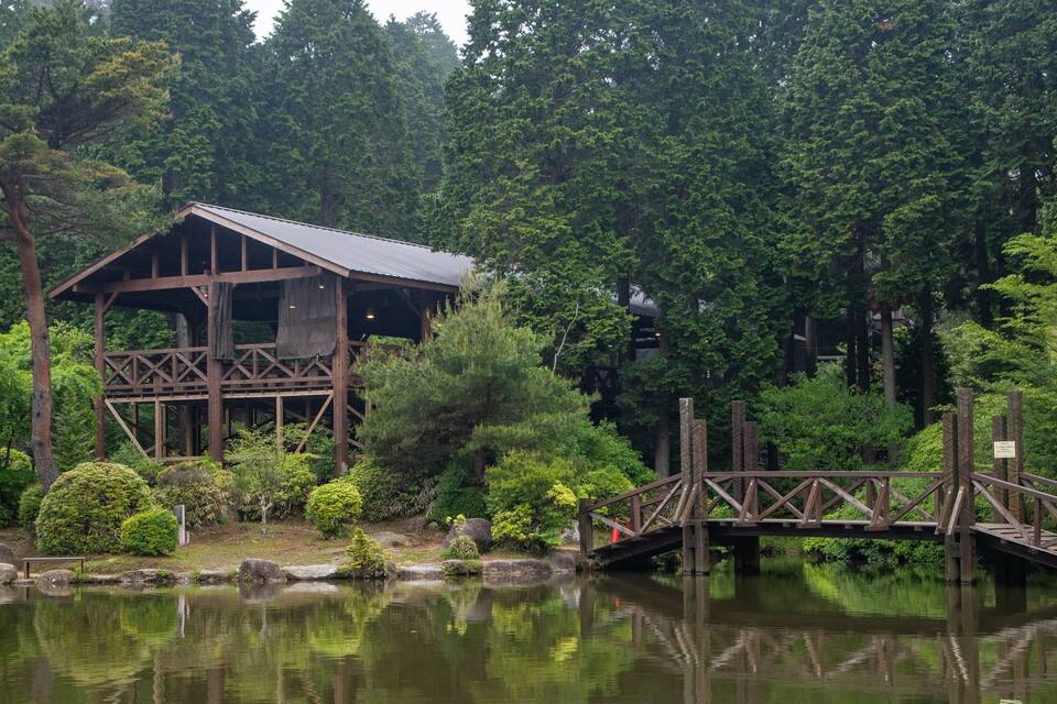 A wooden structure with a metal roof by a calm lake, surrounded by dense trees.
