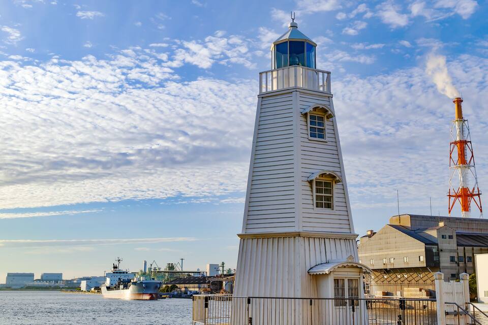 A white lighthouse with a blue top, a boat in the water, and industrial buildings in the background.