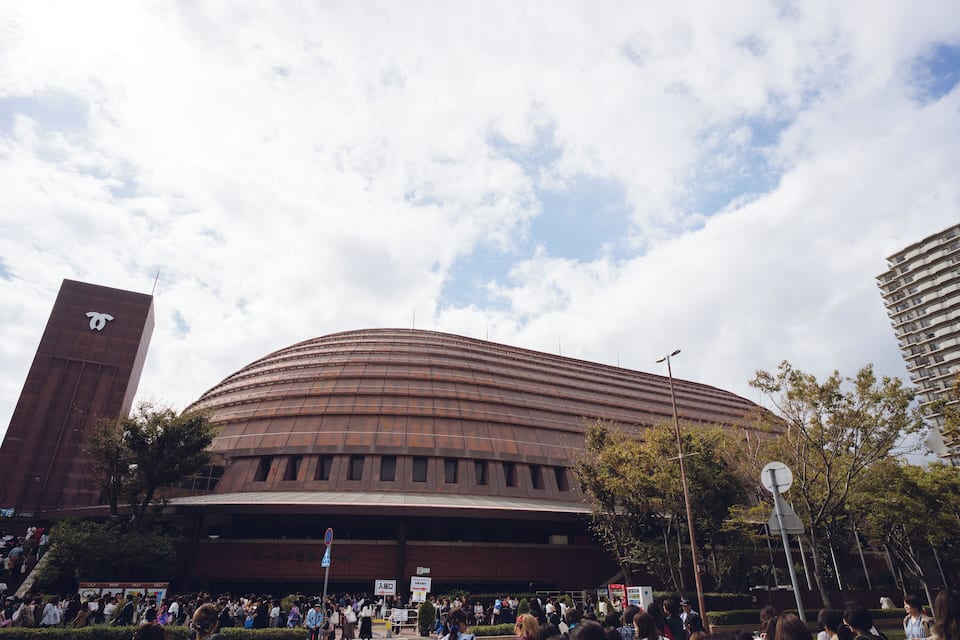 A large, modern stadium with a curved roof, surrounded by trees and tall buildings.