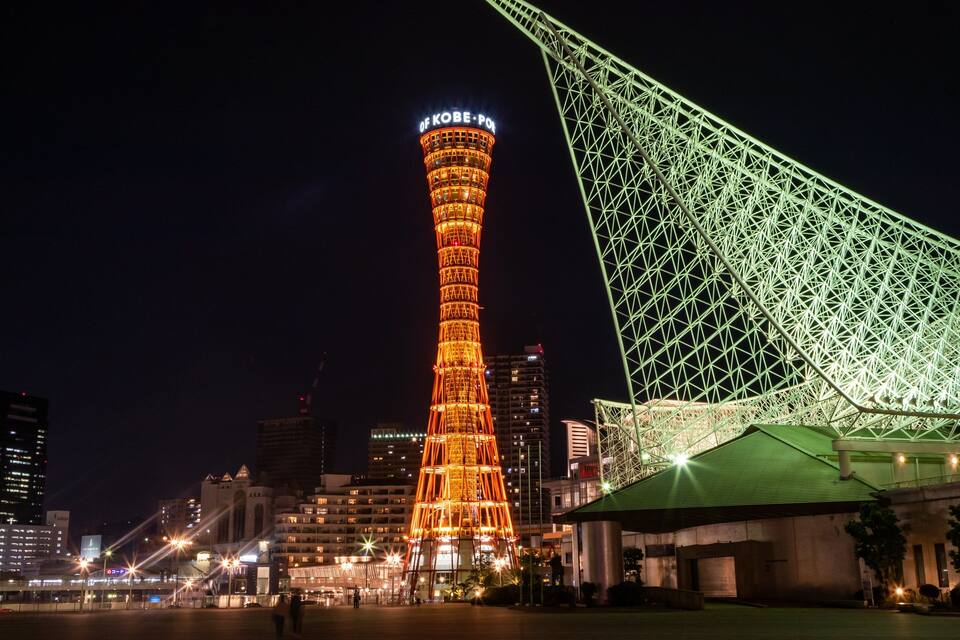 A brightly lit tower with "OF KOBE-PON" on top, a green-lit structure, and surrounding buildings at night.