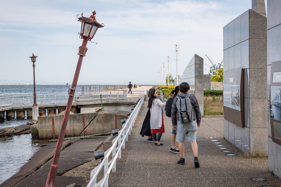 A coastal walkway with a metal lamp post, a concrete barrier, and a stone wall with informational plaques.