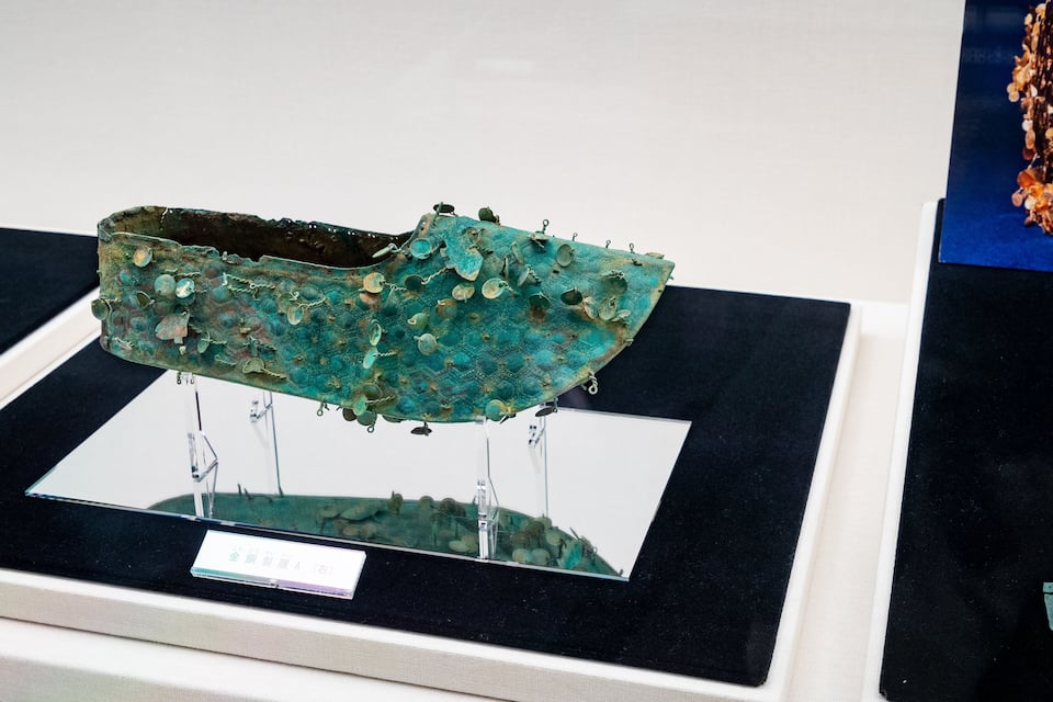 Ein bronzenes Artefakt mit grünem Patina auf einem Podest, versehen mit einer Beschriftung.