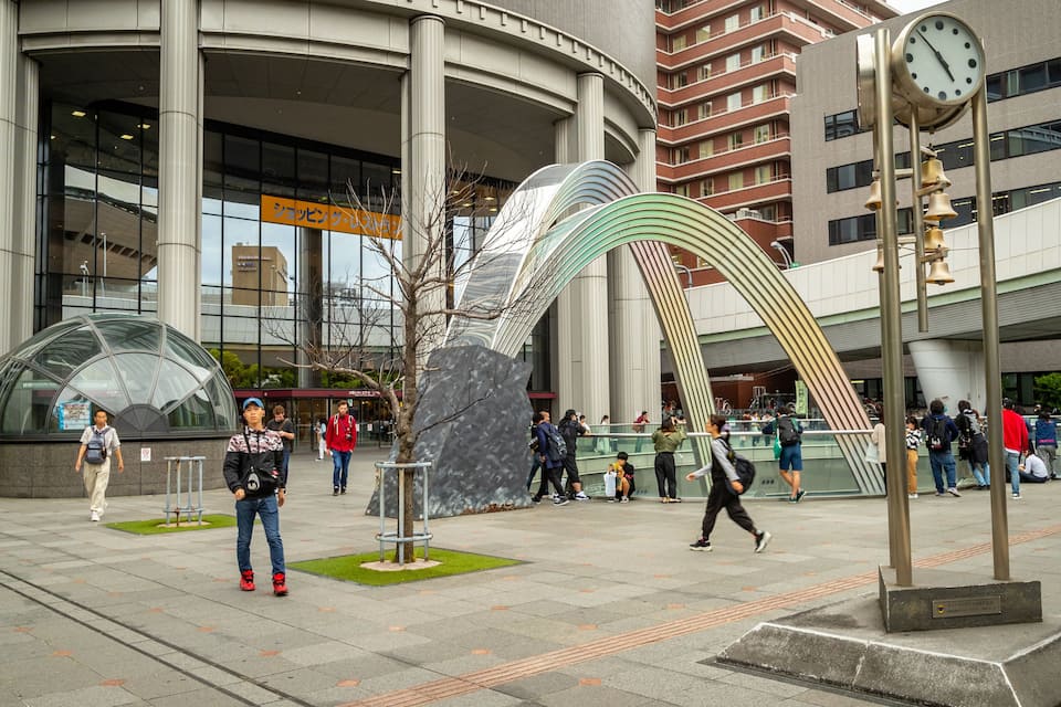 Een modern stadsplein met een groot glazen gebouw, een opvallende metalen sculptuur en een klokkentoren.